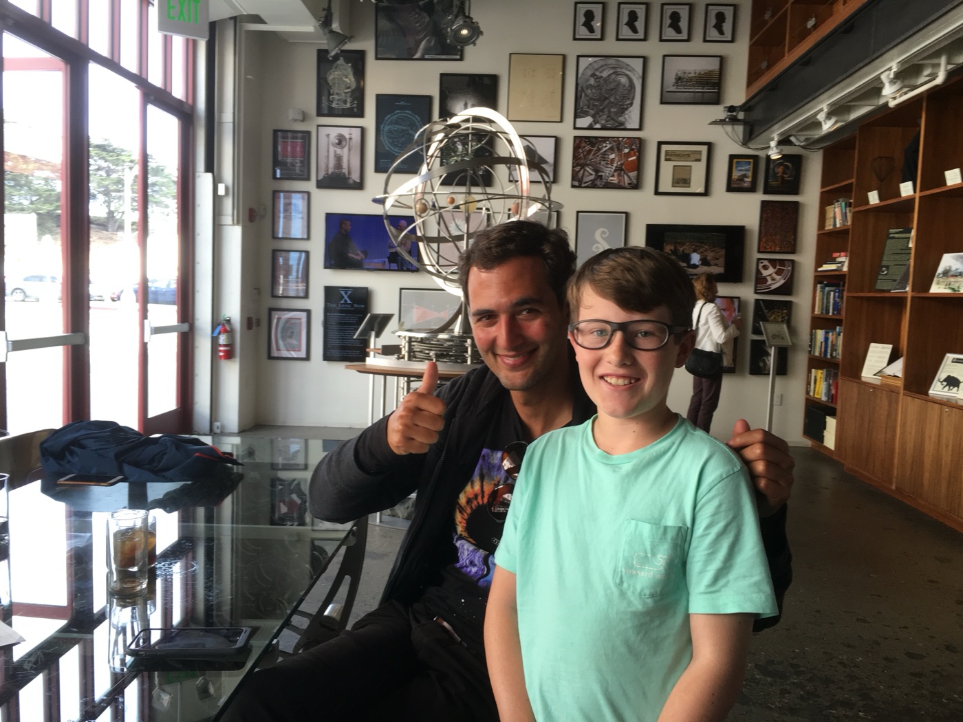 A man giving a thumbs up beside a boy in glasses, with the orrery and a wall of framed prints behind them