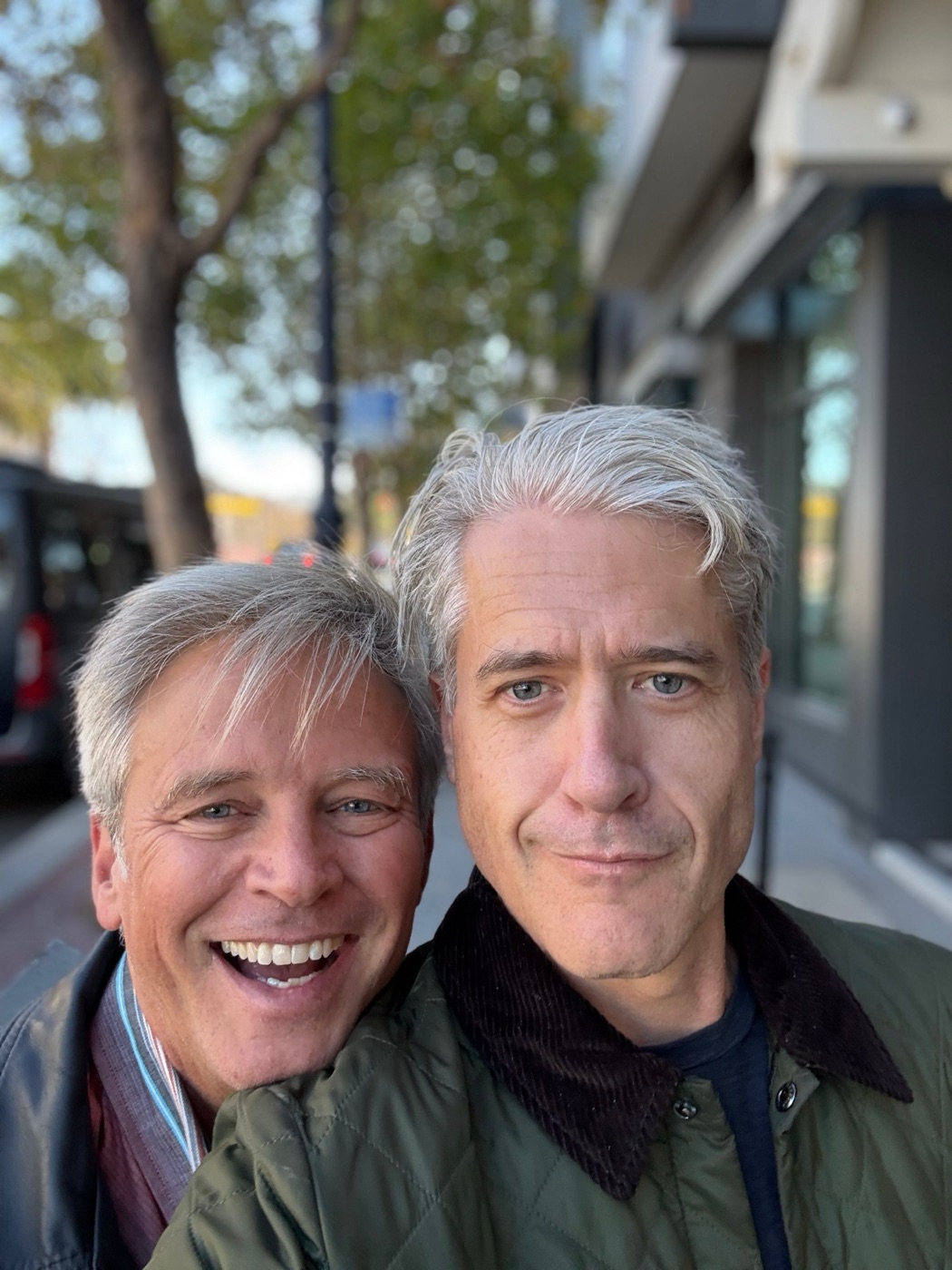 Two gray-haired men smile into the camera on a San Francisco sidewalk